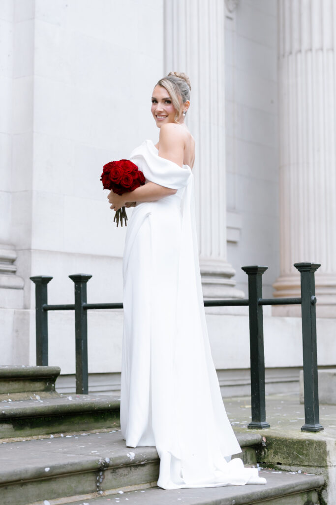 A woman in a white off-shoulder wedding dress stands on stone steps, holding a bouquet of red roses and smiling over her shoulder. She is in front of a white building with tall columns.