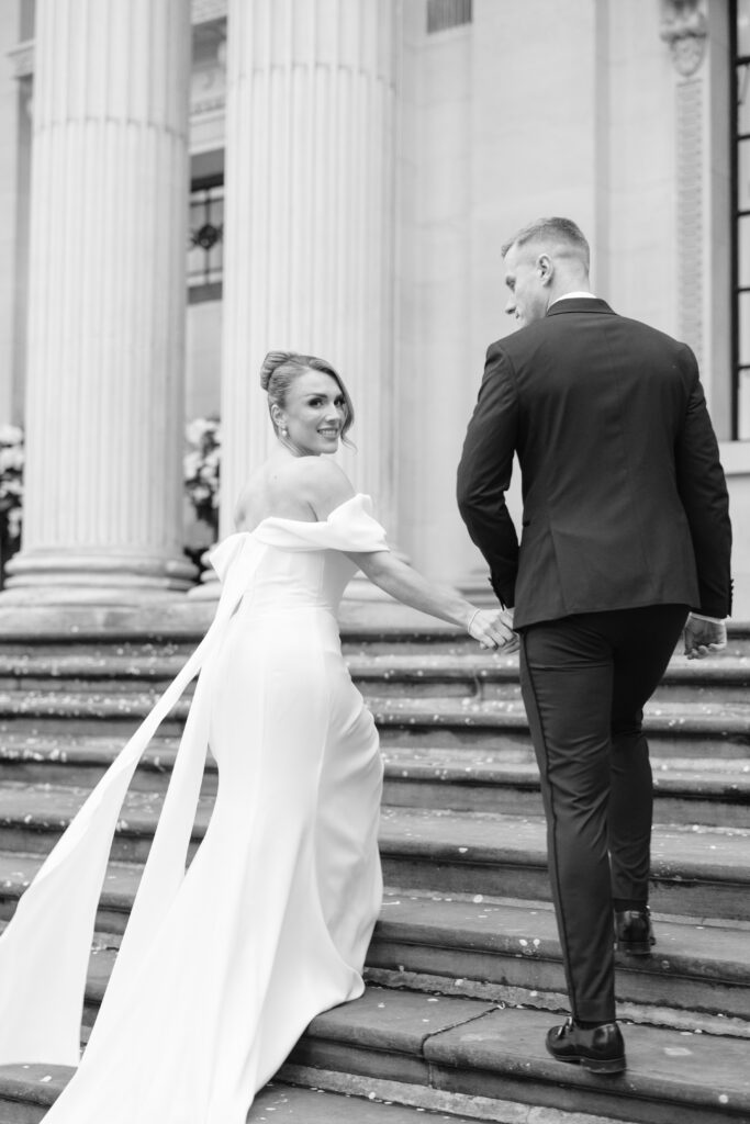 A bride in an off-the-shoulder wedding gown holds hands with a groom in a suit as they walk up stone steps, smiling back, in front of a building with tall columns. Black and white photograph.