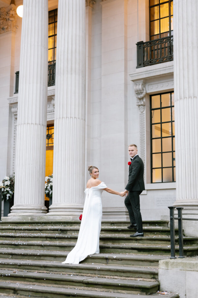 A bride in a white dress and groom in a black suit stand on stone steps, holding hands, in front of a grand building with tall columns and large windows. The bride looks back over her shoulder.
