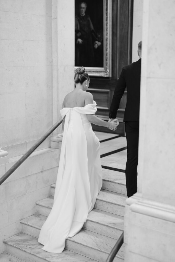 A bride in an off-the-shoulder white gown holds hands with a man in a dark suit as they walk up marble stairs inside an elegant building. A framed painting hangs on the wall above them.