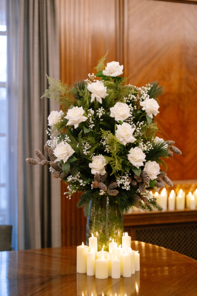 A vase of white roses, green foliage, and pinecones sits on a wooden table with a group of lit white candles in front. In the background, more candles are arranged on a ledge against a wood-paneled wall.
