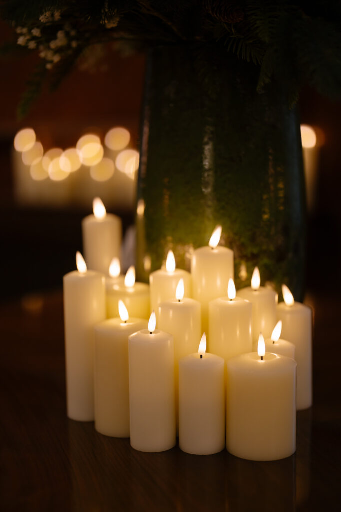 A group of white pillar candles of varying heights are lit and clustered together on a dark surface, creating a warm, soft glow. Blurred lights and greenery are visible in the background.