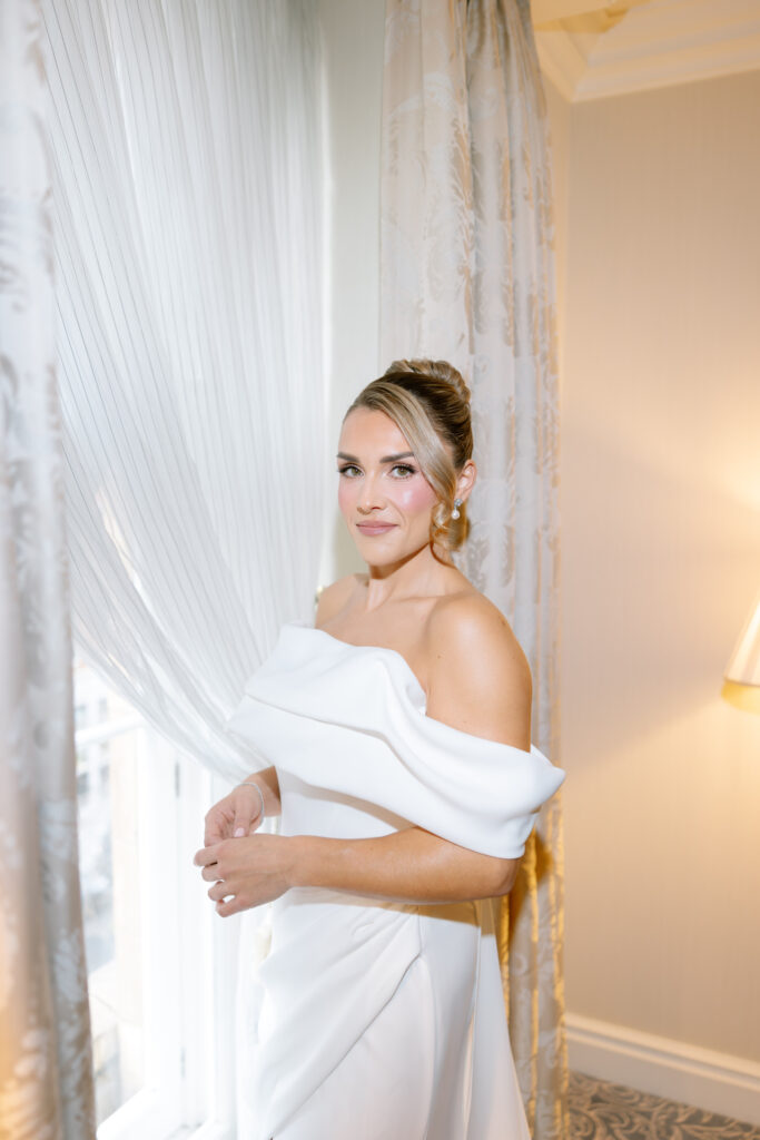 A woman in an elegant off-the-shoulder white dress stands by a window with sheer curtains, her hair styled in an updo, wearing subtle makeup and gold hoop earrings, in a softly lit, neutral-toned room.