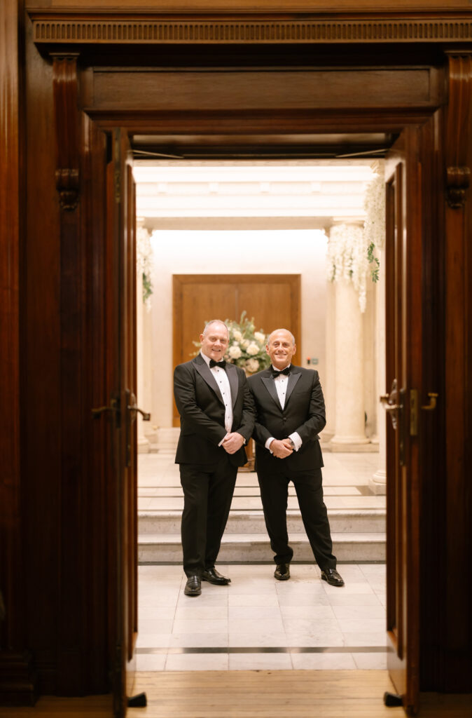Two men in black tuxedos stand side by side, smiling, framed by a large wooden doorway. Behind them is an elegant, well-lit hall with flowers and a wooden double door.