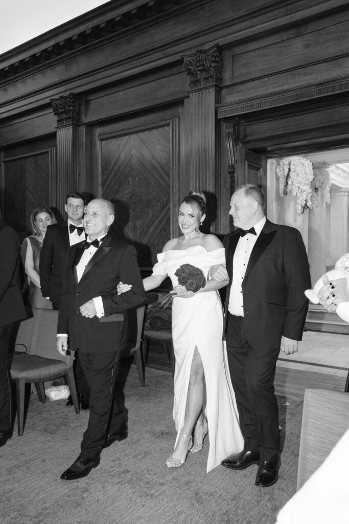 A smiling bride in an off-shoulder wedding dress holds a bouquet and walks down the aisle arm-in-arm with two men in tuxedos, inside an elegant wood-paneled room, as guests look on.