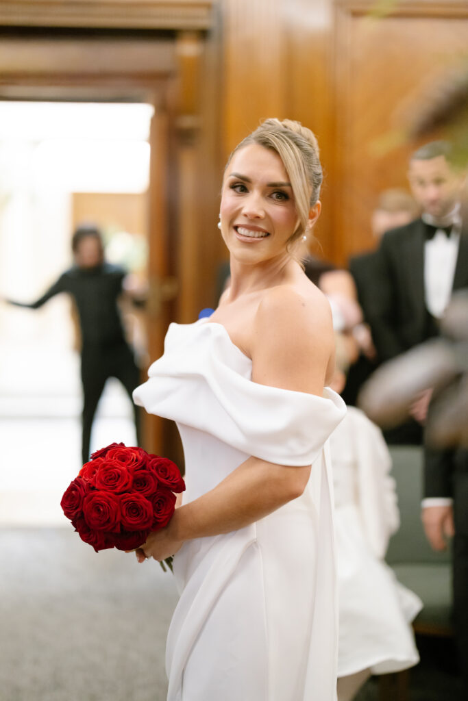 A smiling woman in an off-the-shoulder white wedding dress holds a bouquet of red roses, standing indoors with wooden paneling and blurred people in the background.