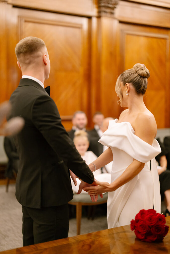 A couple dressed in formal attire stands holding hands during a wedding ceremony in a wood-paneled room, with guests seated in the background and a bouquet of red roses on a table nearby.