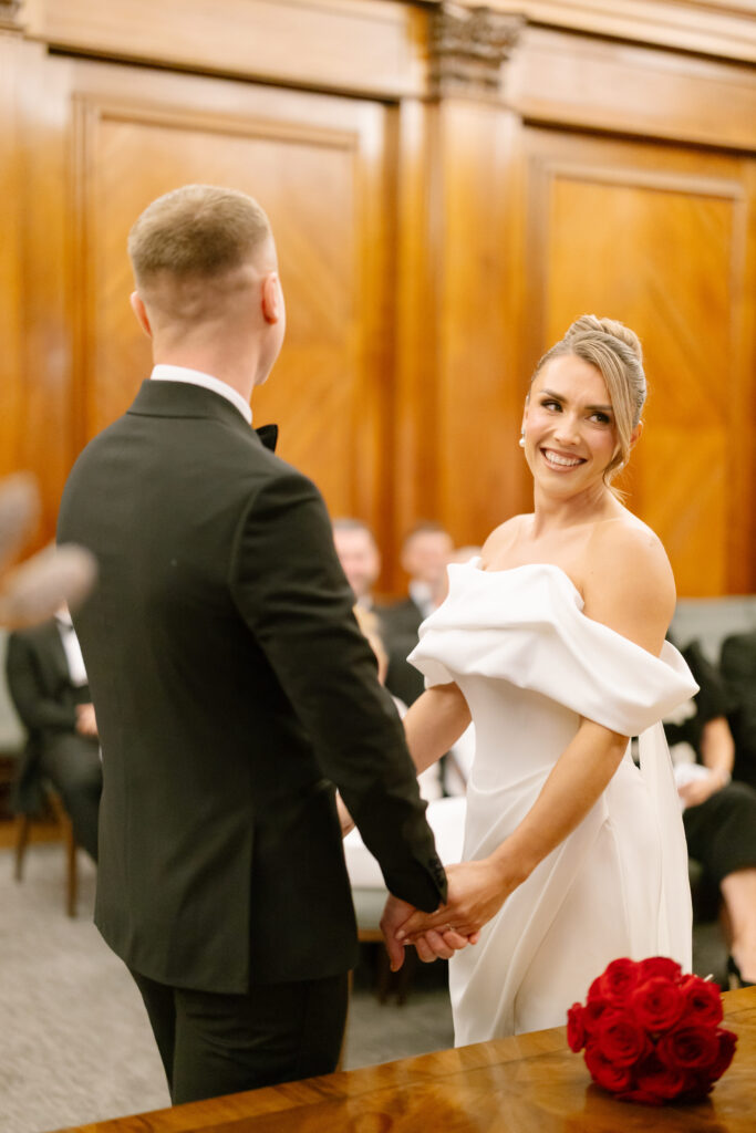 A bride in an off-shoulder white dress smiles while holding hands with the groom in a black suit, standing in a wood-paneled room with seated guests and a bouquet of red roses on a table nearby.