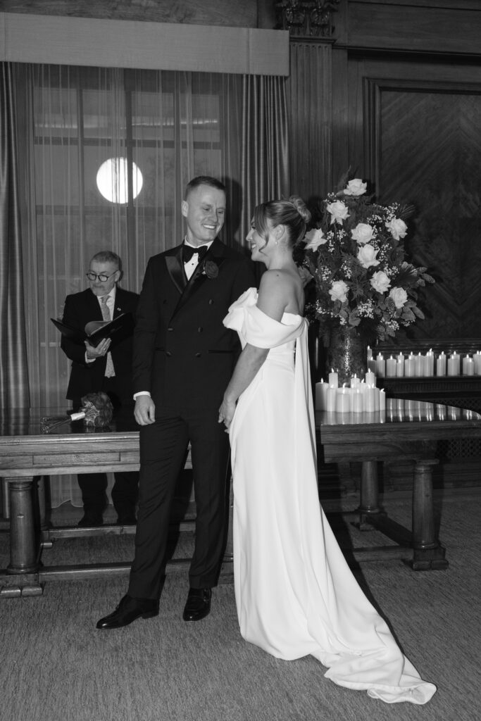 A couple in formal attire stands smiling at each other during their wedding ceremony, with an officiant reading behind them. They are indoors, near a table with a large flower arrangement and dozens of lit candles.