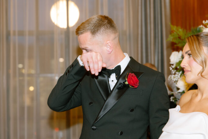 A groom in a black tuxedo with a red rose boutonniere wipes his eyes, appearing emotional, while standing beside a bride in an off-the-shoulder white dress. They are indoors with soft lighting and floral decorations in the background.