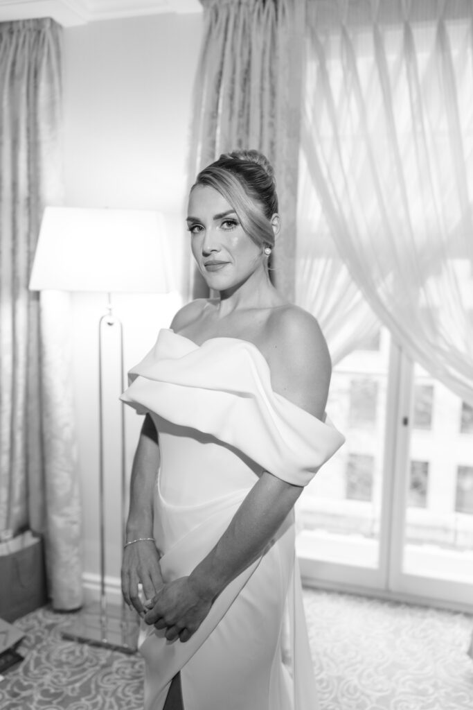 A woman in an elegant off-shoulder gown stands indoors by a window with sheer curtains, smiling softly. The black-and-white photo shows her hair styled in an updo and she wears subtle jewelry.
