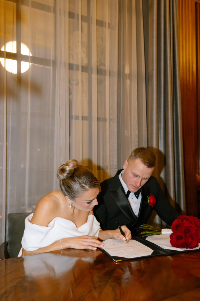 A bride in an off-shoulder white dress signs a document at a wooden table while the groom, in a black tuxedo with a red rose boutonniere, sits beside her. A bouquet of red roses rests on the table.