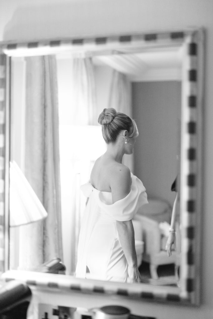 A woman in an off-the-shoulder white dress with her hair in a bun is seen from behind, reflected in a large mirror in a softly lit, elegant room.