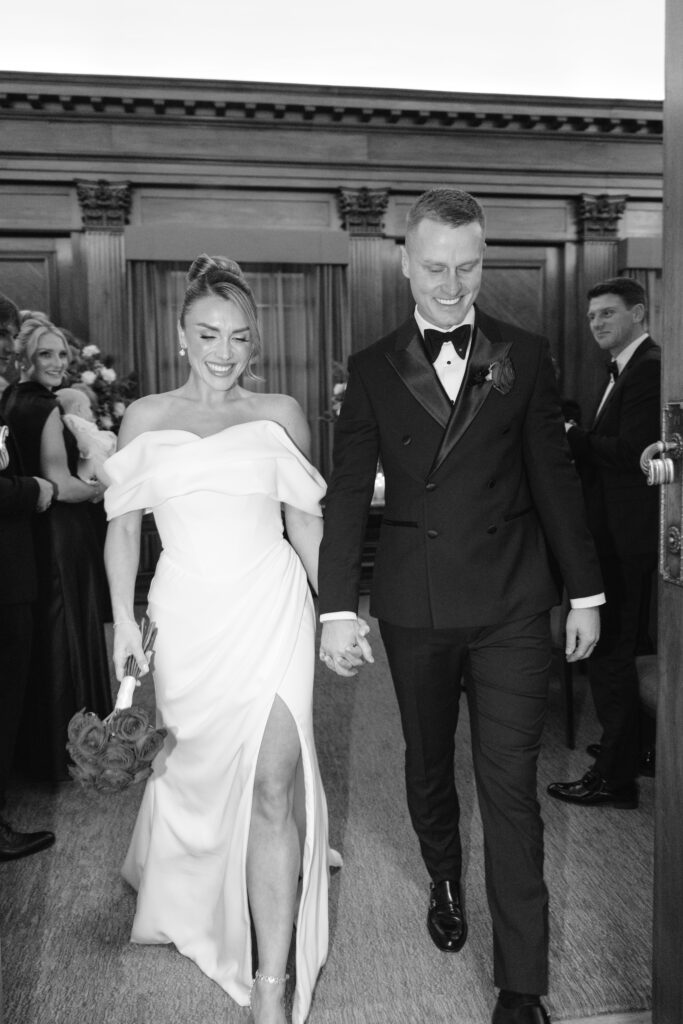 A smiling bride in an off-the-shoulder white dress holds a bouquet of roses and walks hand-in-hand with her groom in a tuxedo, surrounded by cheering guests in formal attire at an indoor wedding.