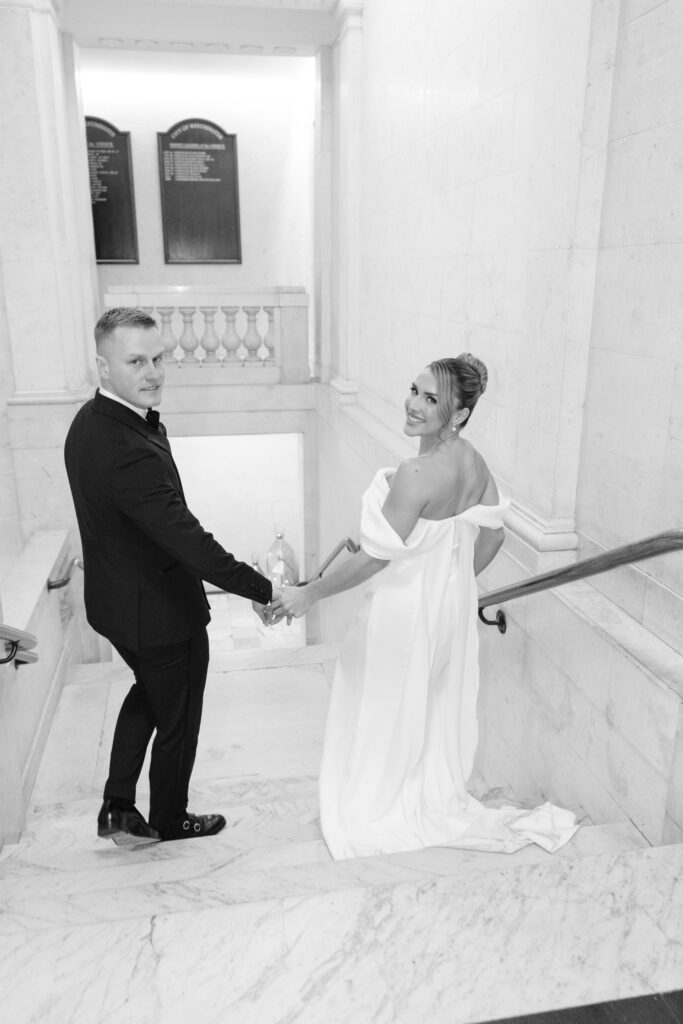 A bride and groom hold hands while walking up marble stairs in a grand building. The bride smiles back at the camera in an off-shoulder gown, while the groom, in a suit, looks over his shoulder. The setting is elegant and bright.
