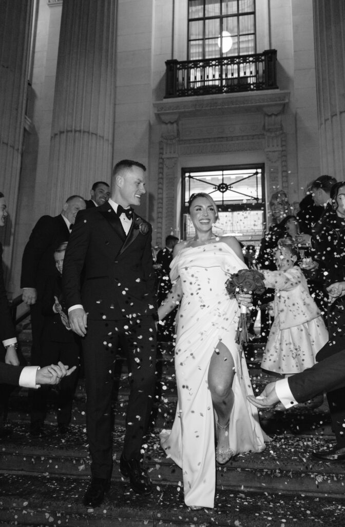 A bride and groom in formal attire walk down steps outside a building as guests throw confetti. The bride smiles, holding a bouquet, while the groom looks at her. The scene is lively and joyful, captured in black and white.