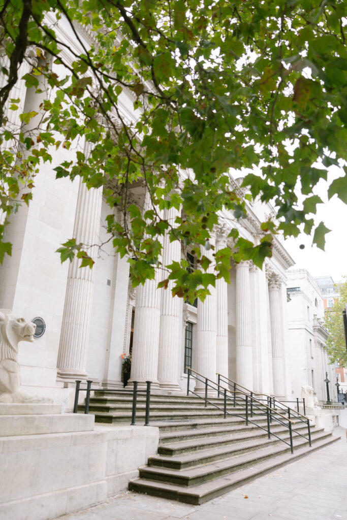 A grand building with tall, white columns and wide stone steps, partially covered by green tree branches. Ornamental sculptures and railings line the entrance.
