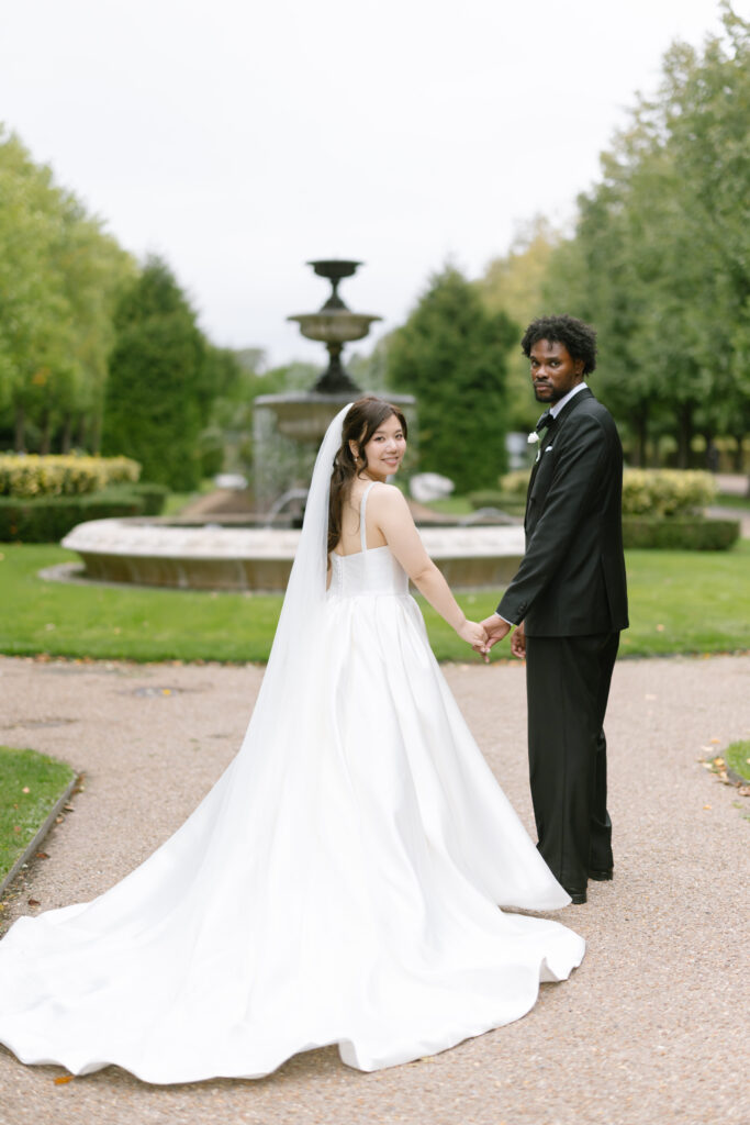 A bride in a white gown and veil holds hands with a groom in a black suit. They stand on a path in front of a fountain, surrounded by green trees, and look back toward the camera, smiling.
