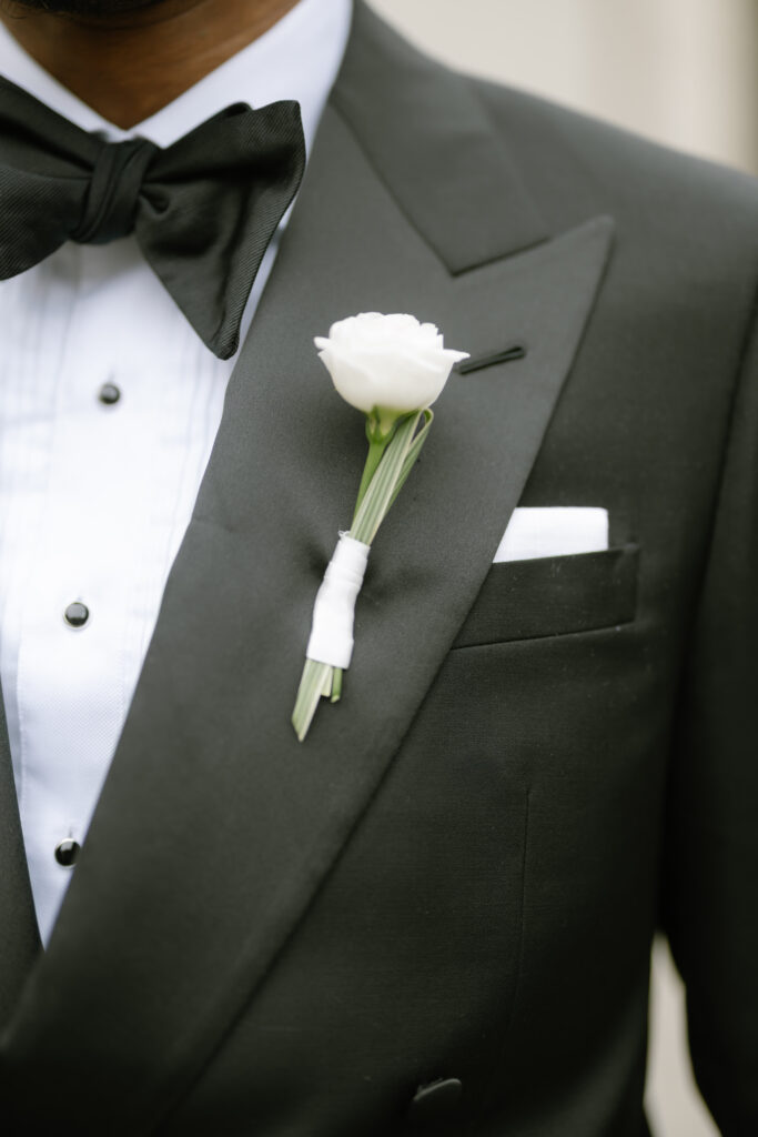 Close-up of a person wearing a black tuxedo with a white dress shirt, black bow tie, and a white boutonnière pinned to the lapel. A white pocket square is visible in the jacket pocket.