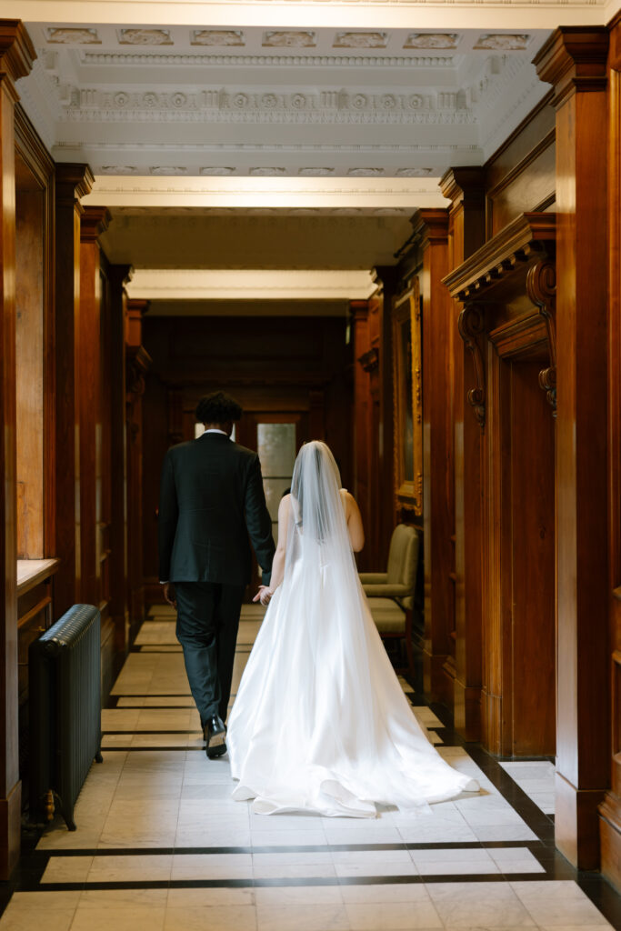 A bride in a white gown and veil walks hand in hand with a groom in a black suit down a hallway lined with wood paneling and ornate molding.