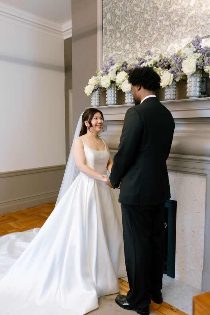 A bride in a white gown and veil holds hands with a groom in a black suit, standing in front of a fireplace decorated with white and purple flowers. They are smiling at each other.