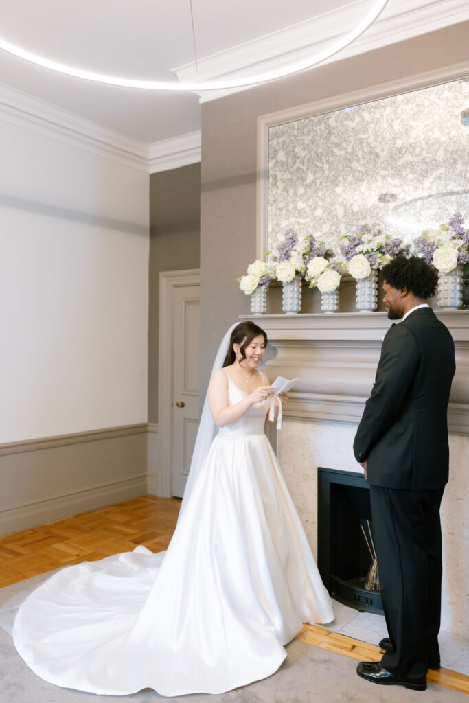 A bride in a white gown reads from a piece of paper while standing beside a groom in a black suit. They are indoors by a fireplace decorated with white and purple flowers. The scene appears joyful and intimate.