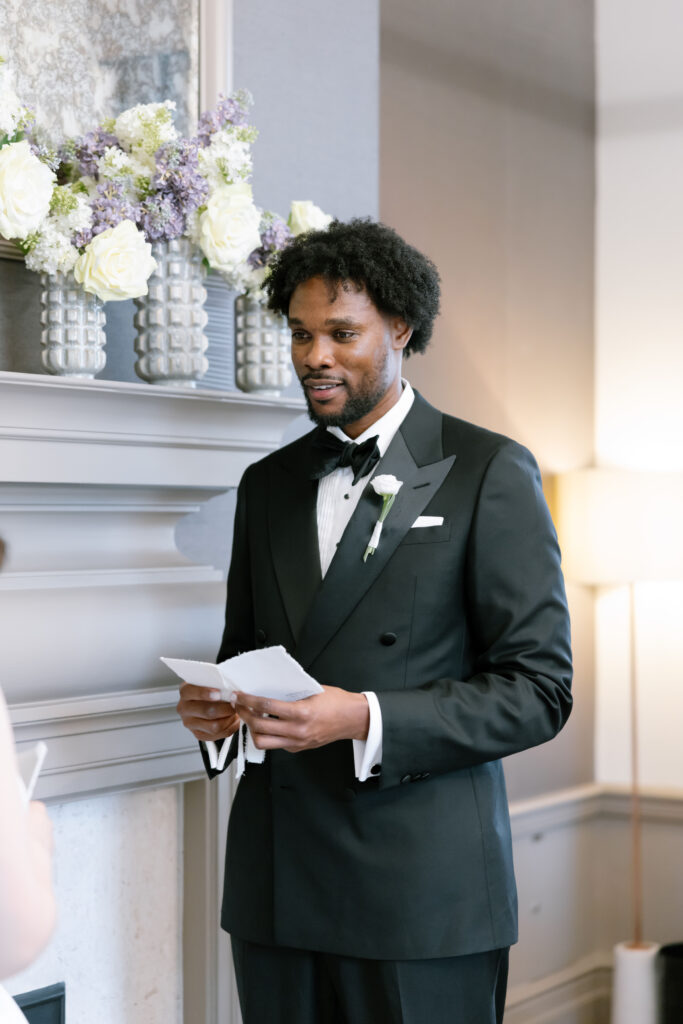 A man in a black tuxedo stands indoors, holding papers and speaking. He is next to a fireplace decorated with vases of white and purple flowers. The setting appears formal, likely during a wedding or ceremony.