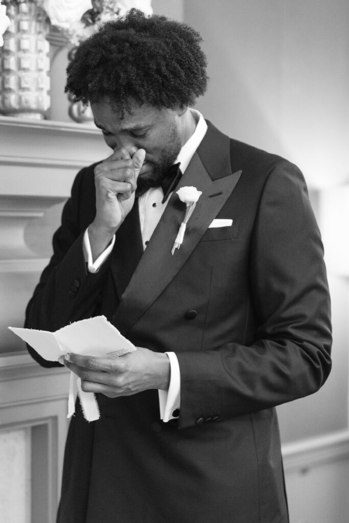 A man in a tuxedo stands indoors, holding a handwritten letter and appearing emotional, covering his mouth with one hand as he reads.