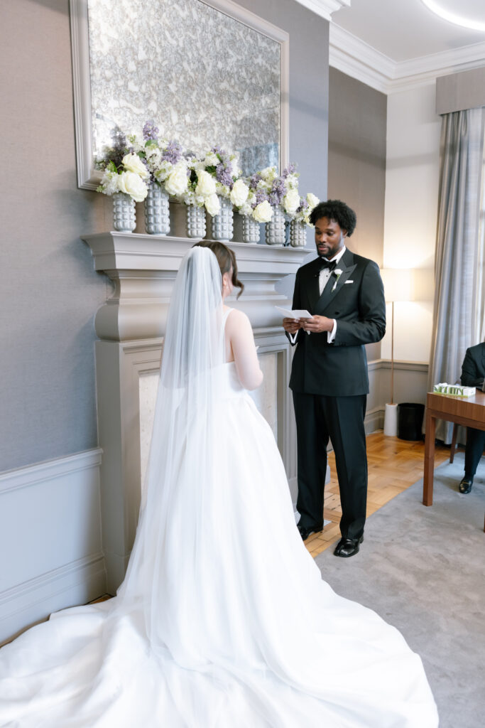 A bride in a white gown and veil stands facing a groom in a black tuxedo, who is reading from a card. They are indoors by a decorated mantel with vases of white and purple flowers.