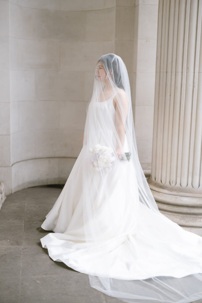 A bride stands indoors near a large column, wearing a long white wedding gown and veil, and holding a bouquet of light-colored flowers. Soft natural light fills the elegant, classical space.