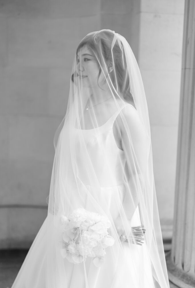 A bride in a white dress and veil holds a bouquet of flowers, standing indoors near a wall, looking to the side and smiling softly. The image is in black and white.