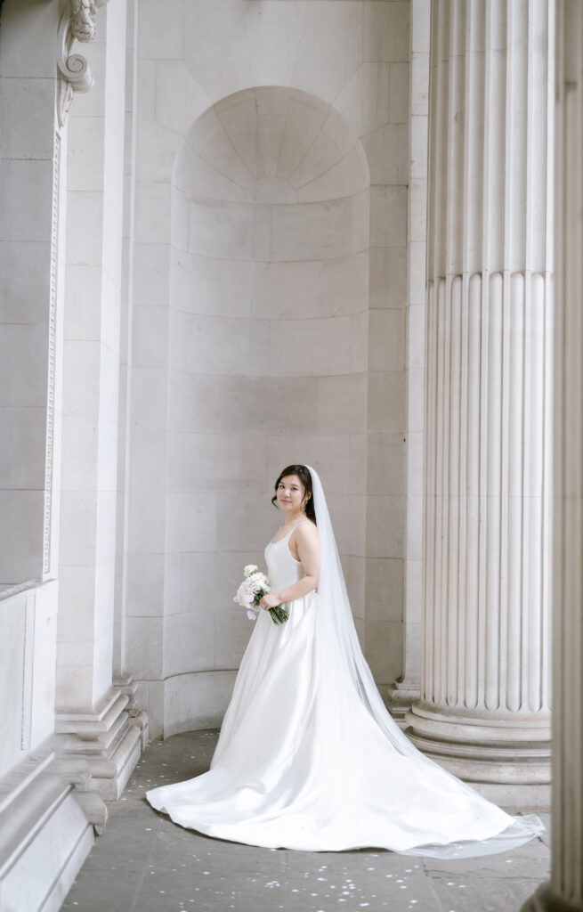 A bride in a white dress and veil stands holding a bouquet of flowers between tall marble columns, smiling softly in an elegant, bright architectural setting.