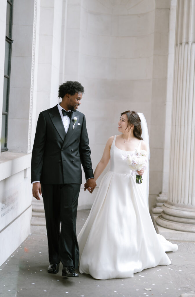 A bride in a white gown and veil holds a bouquet and smiles at the groom in a black tuxedo as they walk hand in hand in a grand, columned hallway.