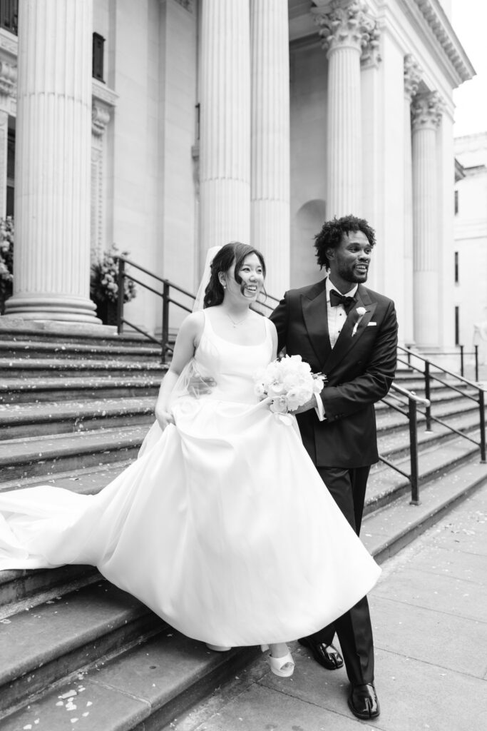 A bride in a flowing white gown and a groom in a black tuxedo smile as they walk down stone steps outside a grand building with tall columns.