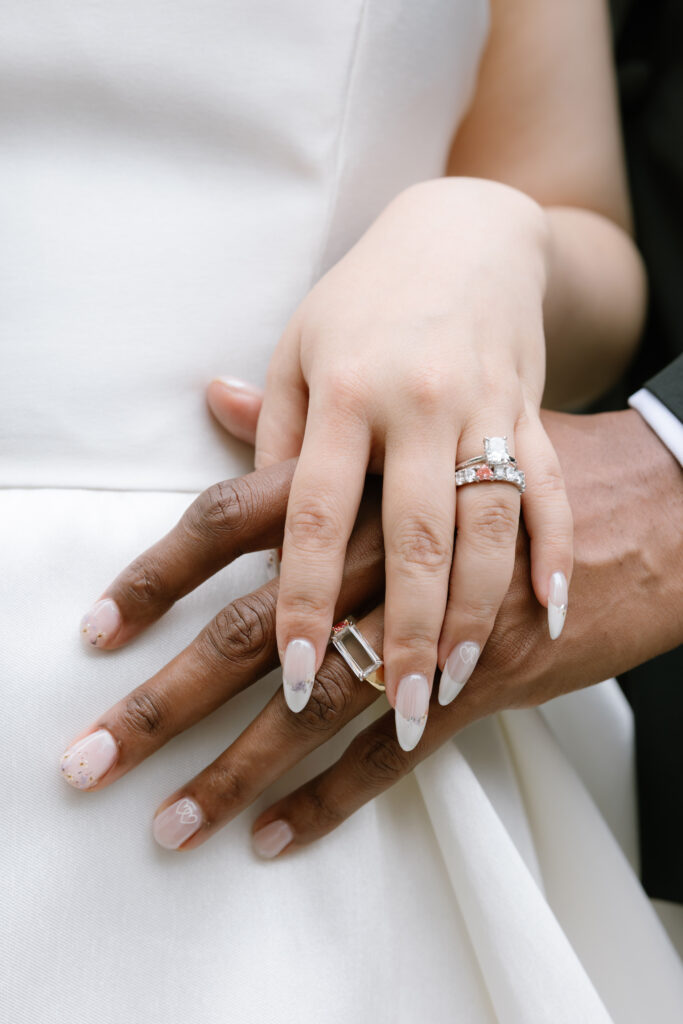 Close-up of a couple’s hands resting on each other, both wearing wedding rings. The bride’s hand is on top, showing a diamond engagement ring and delicate nail art. The groom’s hand is beneath, also wearing a band.