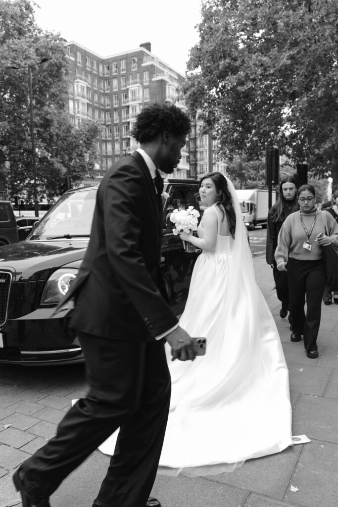 A bride in a white dress holding a bouquet stands near a parked car on a city street, while a man in a suit walks past her and two women stand nearby. Trees and tall buildings are visible in the background.