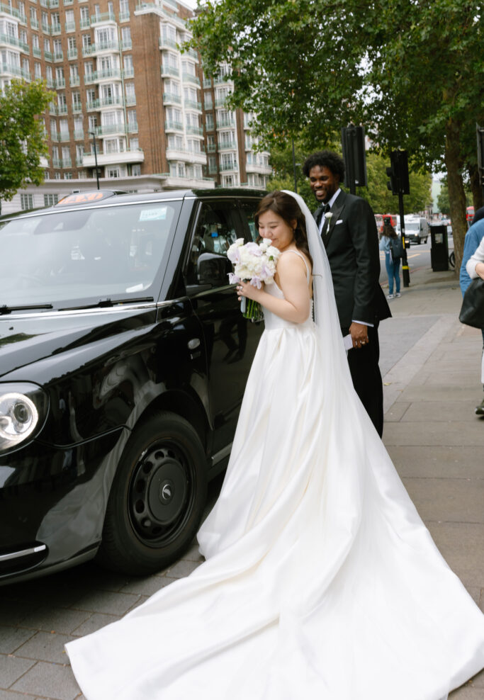A bride in a white gown holding a bouquet stands beside a black taxi on a city street, smiling, with a groom in a black suit behind her. Tall buildings and trees are visible in the background.