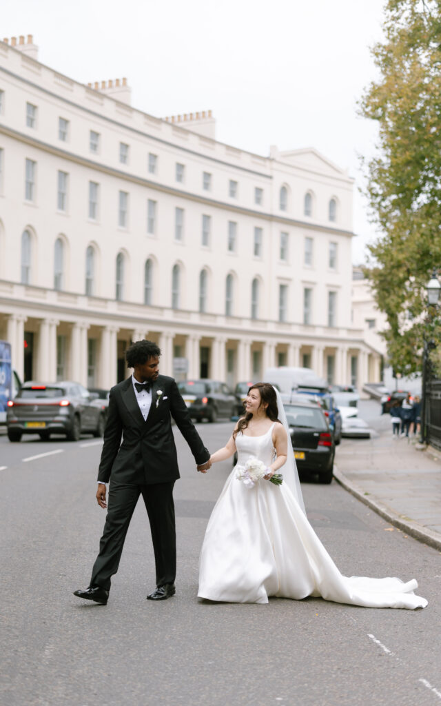 A bride in a white gown and a groom in a black tuxedo hold hands and smile while walking on a city street lined with white buildings and parked cars.