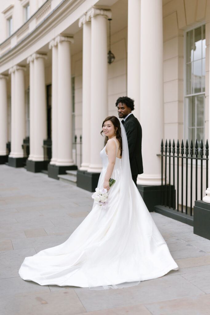 A bride in a white gown holds a bouquet and smiles at the camera while standing next to a groom in a dark suit. They are outside a building with tall columns and black railings.