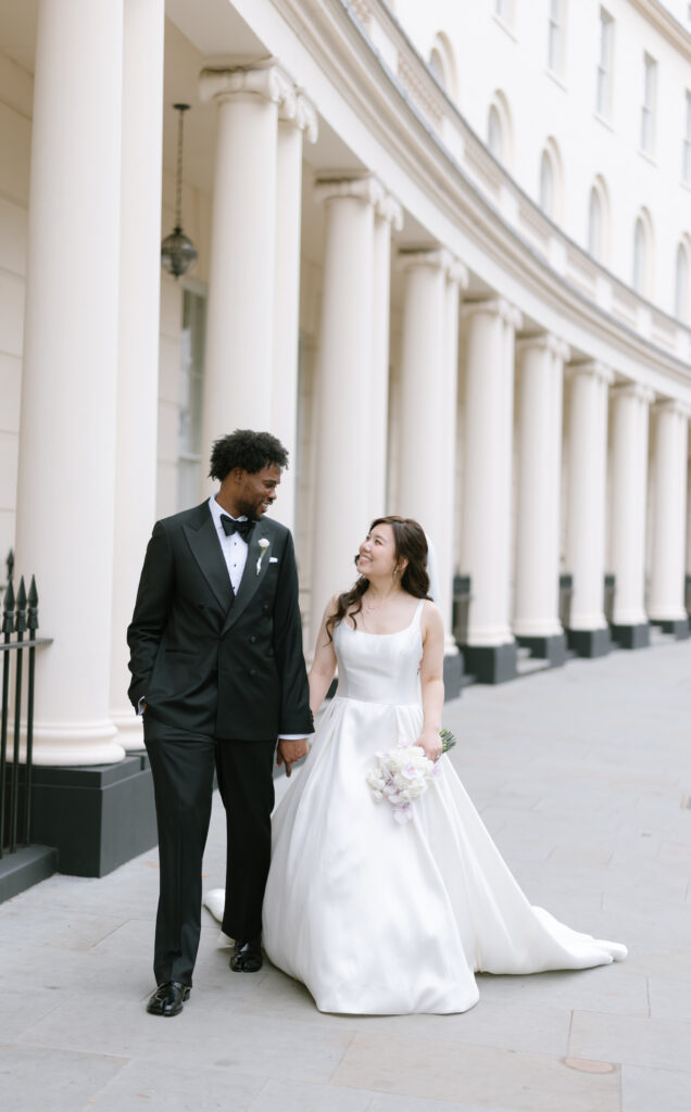 A bride in a white gown and a groom in a black tuxedo walk hand in hand, smiling at each other, outside a grand building with tall white columns.
