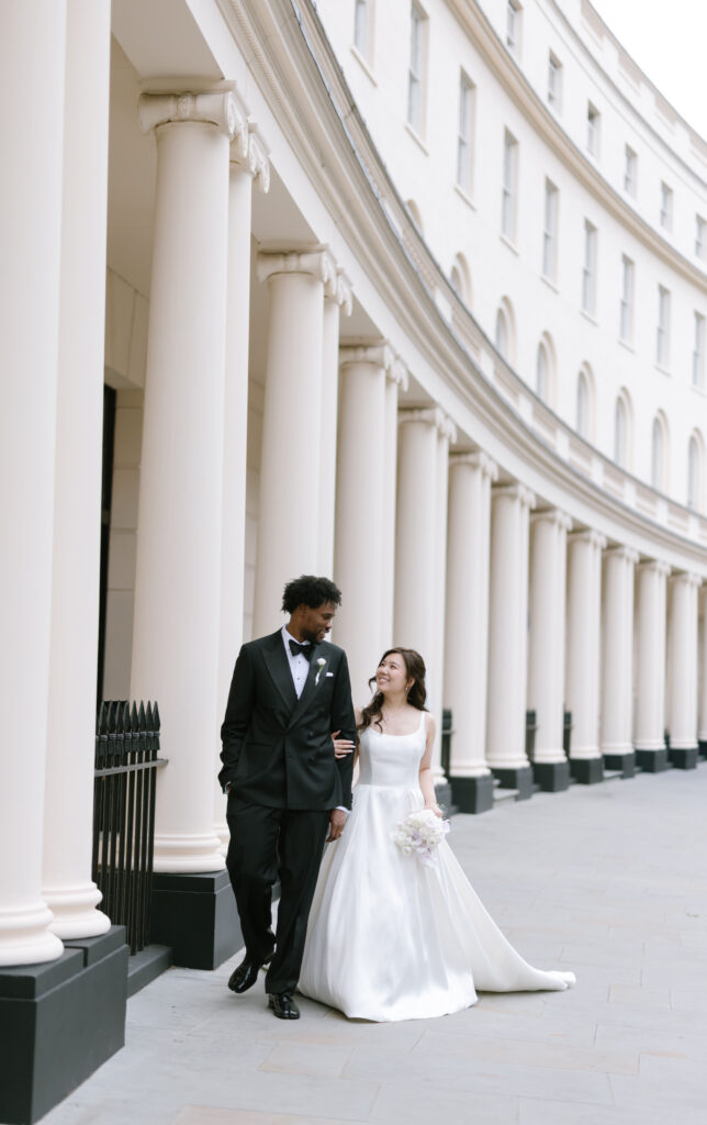 A bride in a white gown and groom in a black tuxedo walk arm in arm, smiling at each other, in front of a curved building with tall columns.