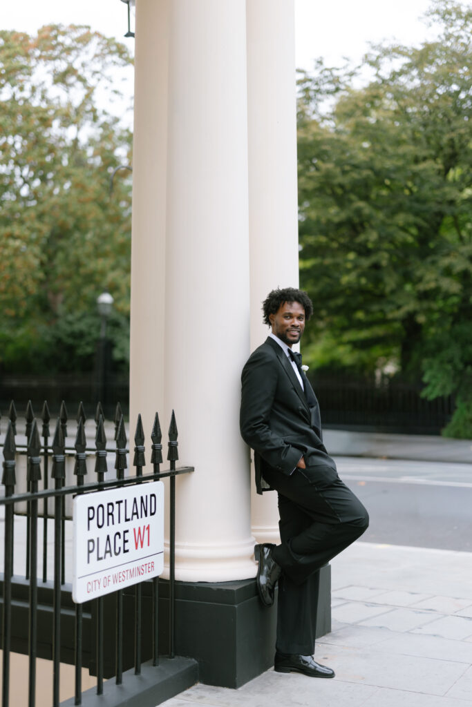 A man in a black tuxedo leans against a white column on a city sidewalk near a Portland Place W1, City of Westminster street sign. Trees and a street are visible in the background.