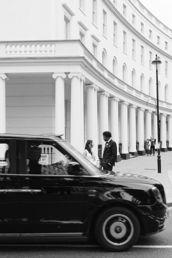 A black taxi drives past a man and woman talking on a city sidewalk in front of a white building with tall columns; the scene is captured in black and white.