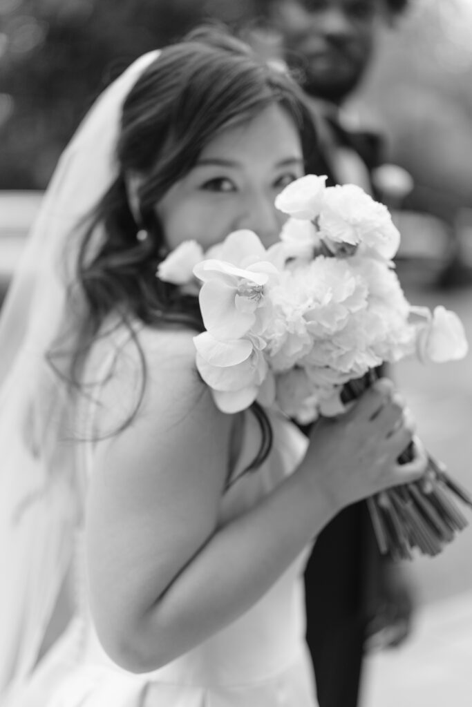 A bride in a white dress holds a bouquet of flowers close to her face and smiles at the camera. An out-of-focus man in a suit stands behind her. The image is in black and white.