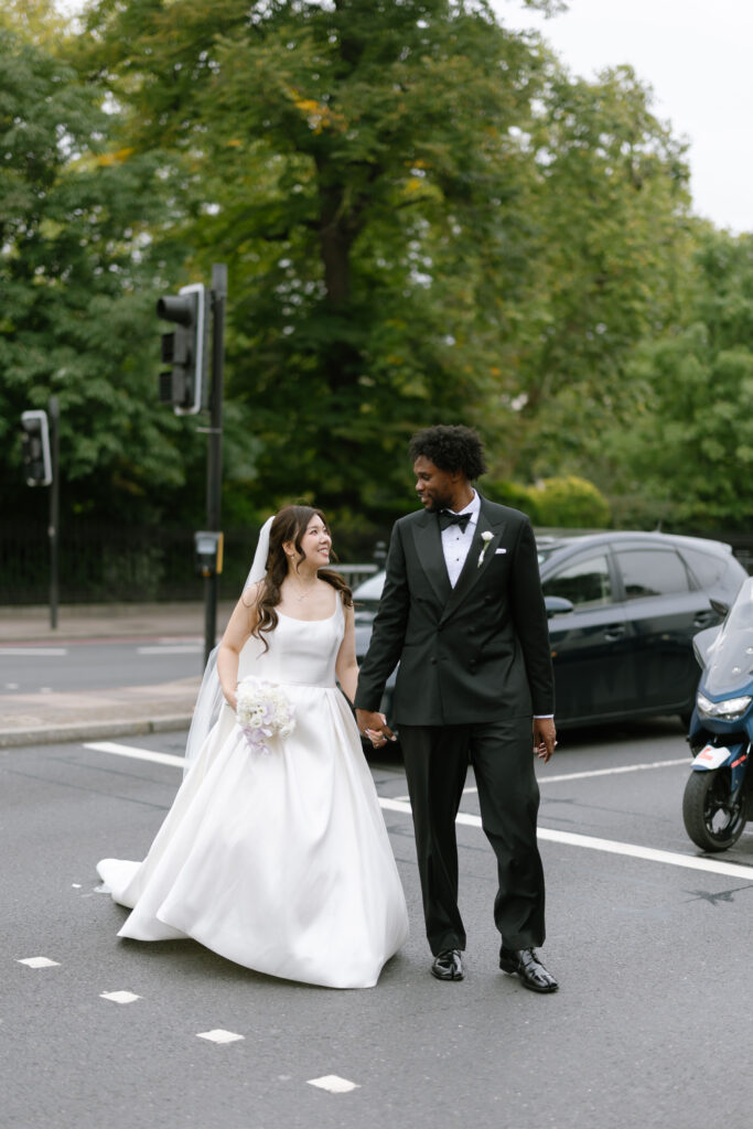 A bride in a white gown and a groom in a black tuxedo hold hands and smile at each other while crossing a street, with trees, a car, and a motorcycle in the background.