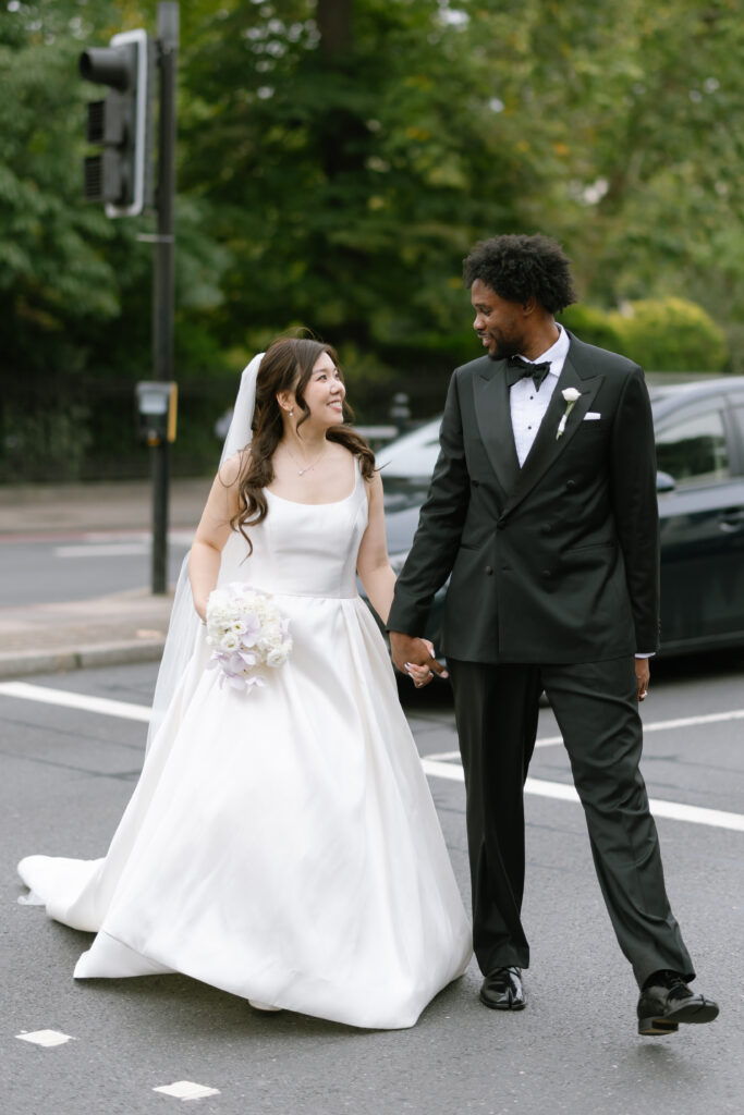A bride in a white gown holding a bouquet and a groom in a black tuxedo hold hands and smile at each other while crossing a street, with greenery and a parked car in the background.