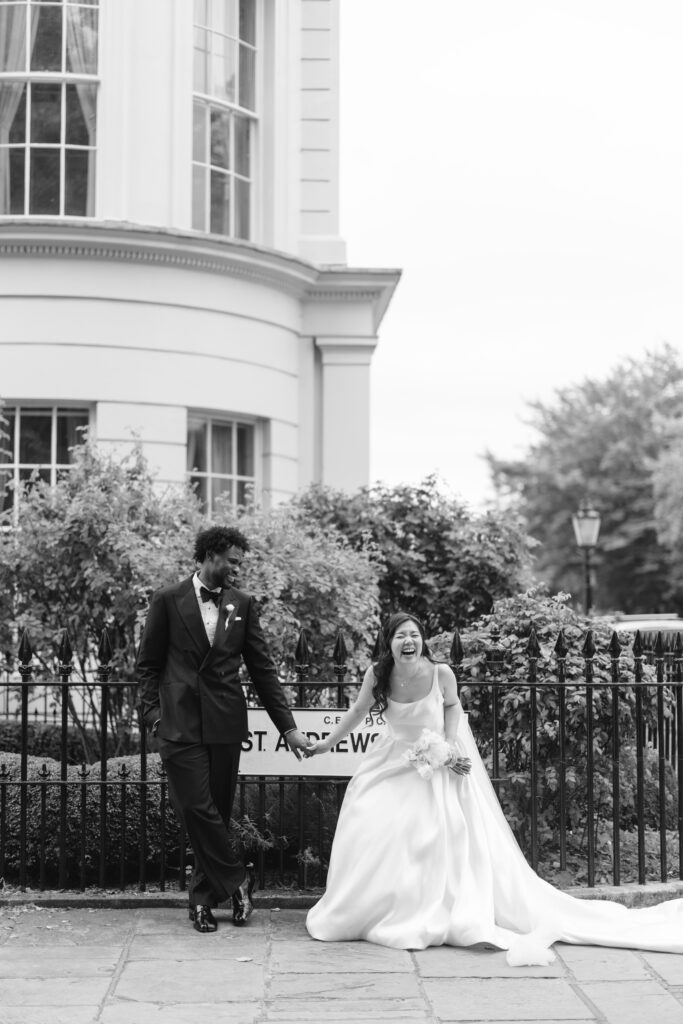 A bride and groom in formal wedding attire stand and laugh together by a wrought-iron fence, with a building and greenery in the background. The scene is in black and white.