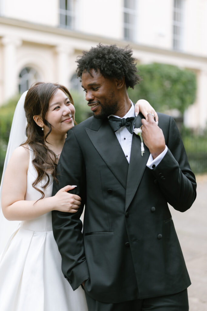 A bride in a white dress and veil embraces a groom in a black tuxedo outdoors. They gaze at each other and smile, with greenery and a building in the background.