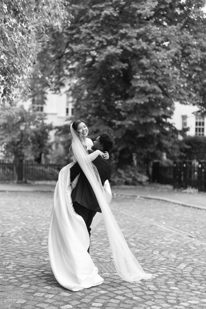 A bride in a long, flowing gown and veil smiles as she is lifted by a person in a suit on a cobblestone street, with trees and buildings in the background. The image is in black and white.