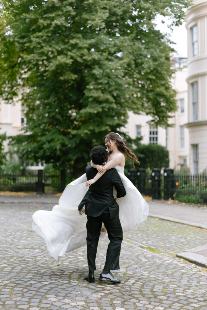 A groom in a black suit spins his bride, who is wearing a flowing white dress, on a cobblestone path outdoors, with large green trees and white buildings in the background. Both appear joyful and carefree.
